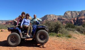 Four-wheel drive vehicle navigating a rocky, dusty trail in the Arizona desert, surrounded by cacti and red rock formations under a bright blue sky.