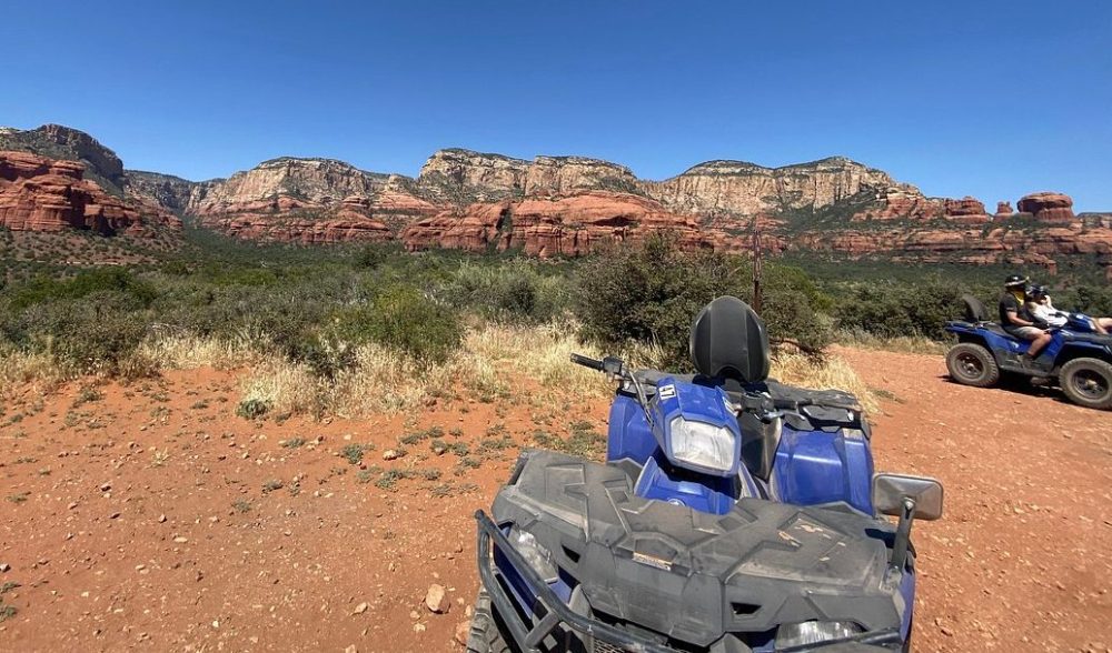 rugged all-terrain vehicle parked on a sandy desert trail, surrounded by towering red rock formations under a bright blue sky, with cacti and sparse desert vegetation in the foreground, showcasing the adventurous spirit of outdoor exploration in Arizona.