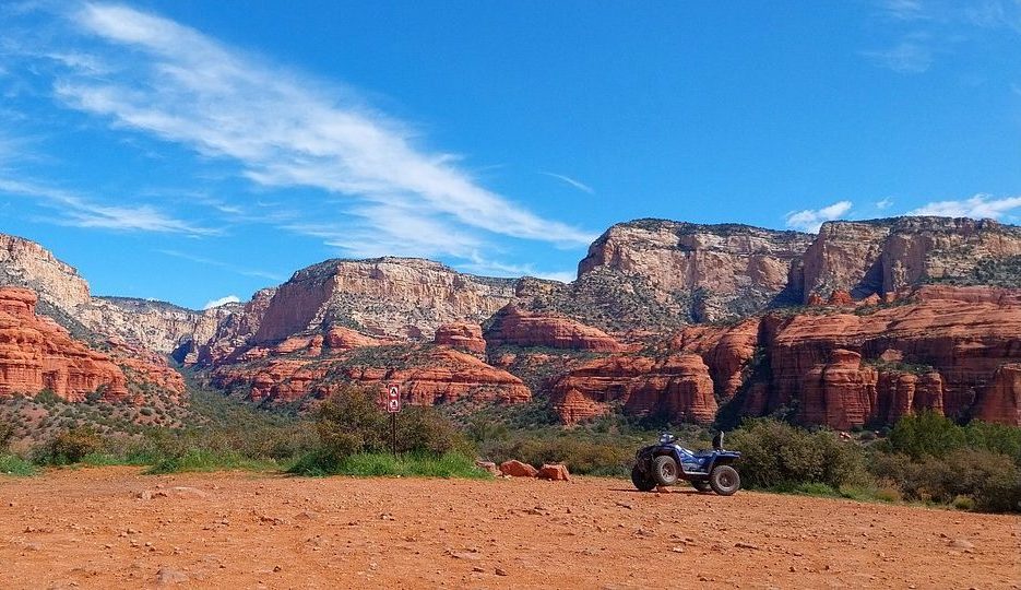 A vibrant outdoor scene showcasing a line of colorful UTVs and ATVs parked along a rugged trail, surrounded by lush green forests and rolling hills under a bright blue sky. Sunlight filters through the trees, casting playful shadows, while some vehicles are partly covered in dirt, hinting at adventures just taken.