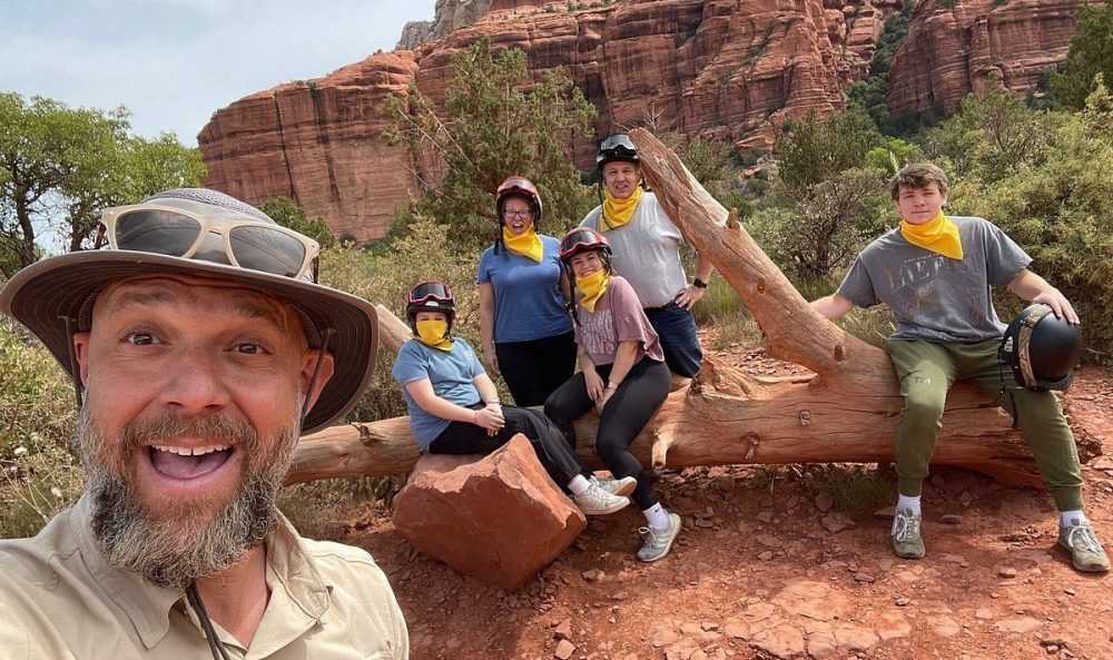 A stunning landscape of Sedona's red rock formations, with rugged terrain and vibrant desert flora, showcasing a lineup of adventurers ready for exploration. The sun sets in the background, casting warm golden hues over the scene, highlighting the adventure awaiting in this picturesque setting.